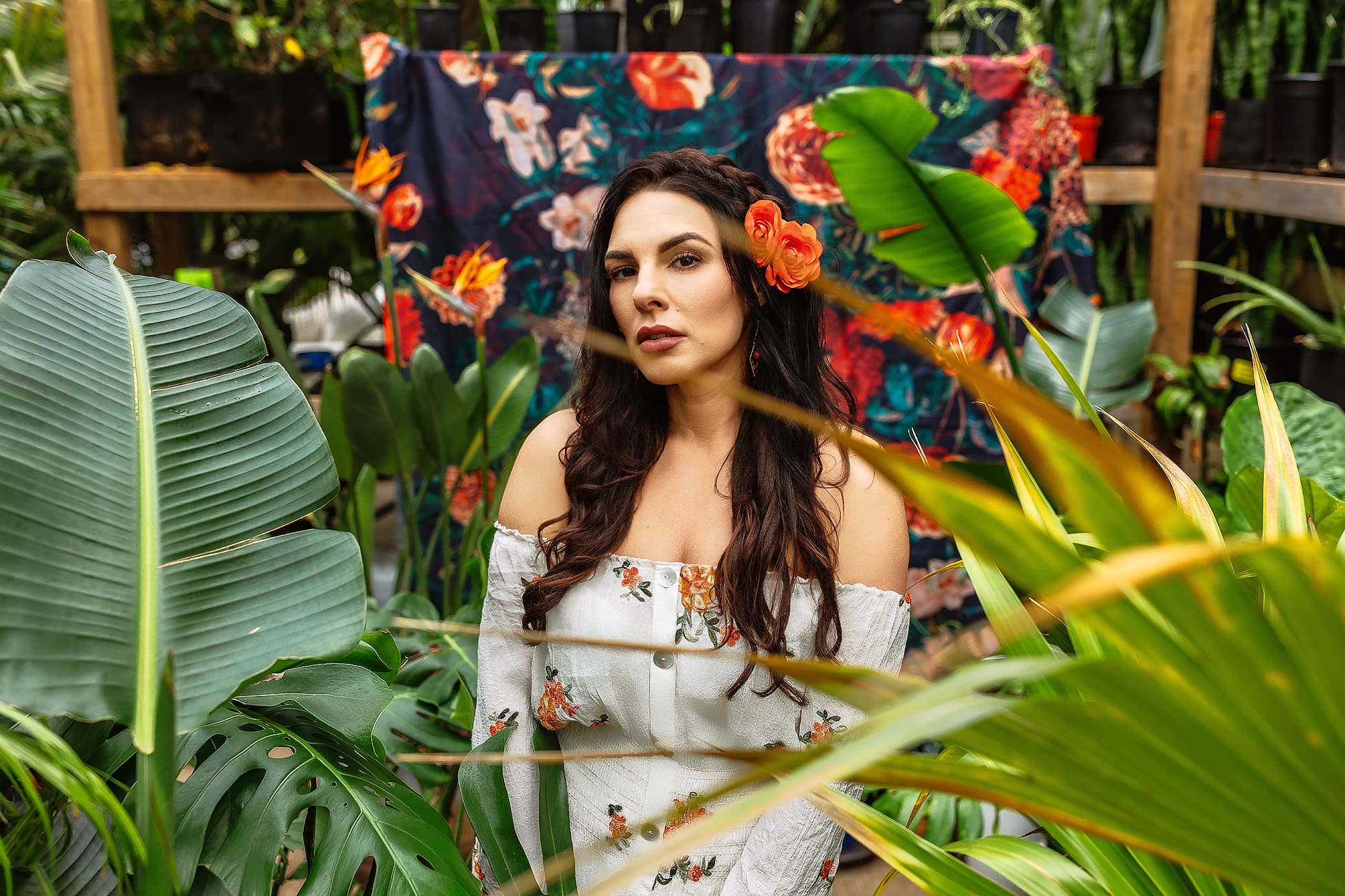 A woman with long, dark hair adorned by an orange flower poses in a floral off-shoulder top amidst lush greenery. Captured in a vibrant, tropical-themed greenhouse setting, this Connecticut headshot exudes natural elegance.