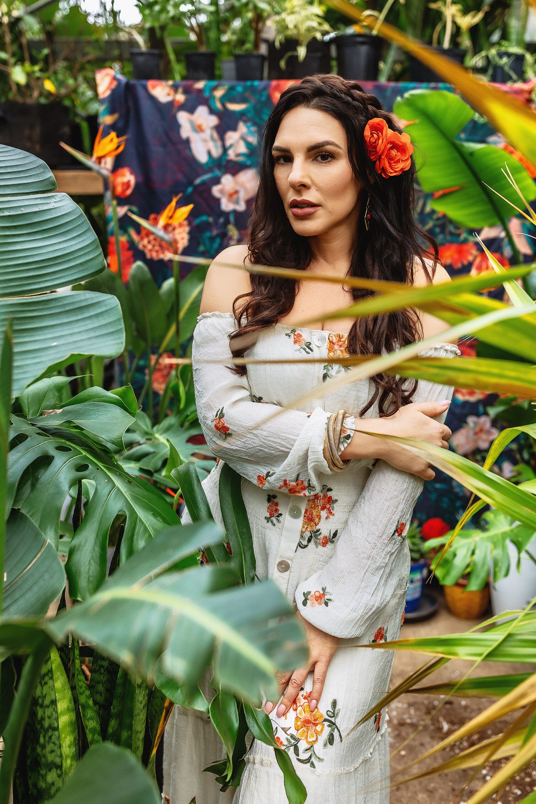 A woman in an off-shoulder floral dress and a red flower in her hair stands gracefully among lush, green plants. Captured by a Connecticut headshot photographer, the vibrant scene is accentuated by a colorful floral backdrop partially visible behind her.