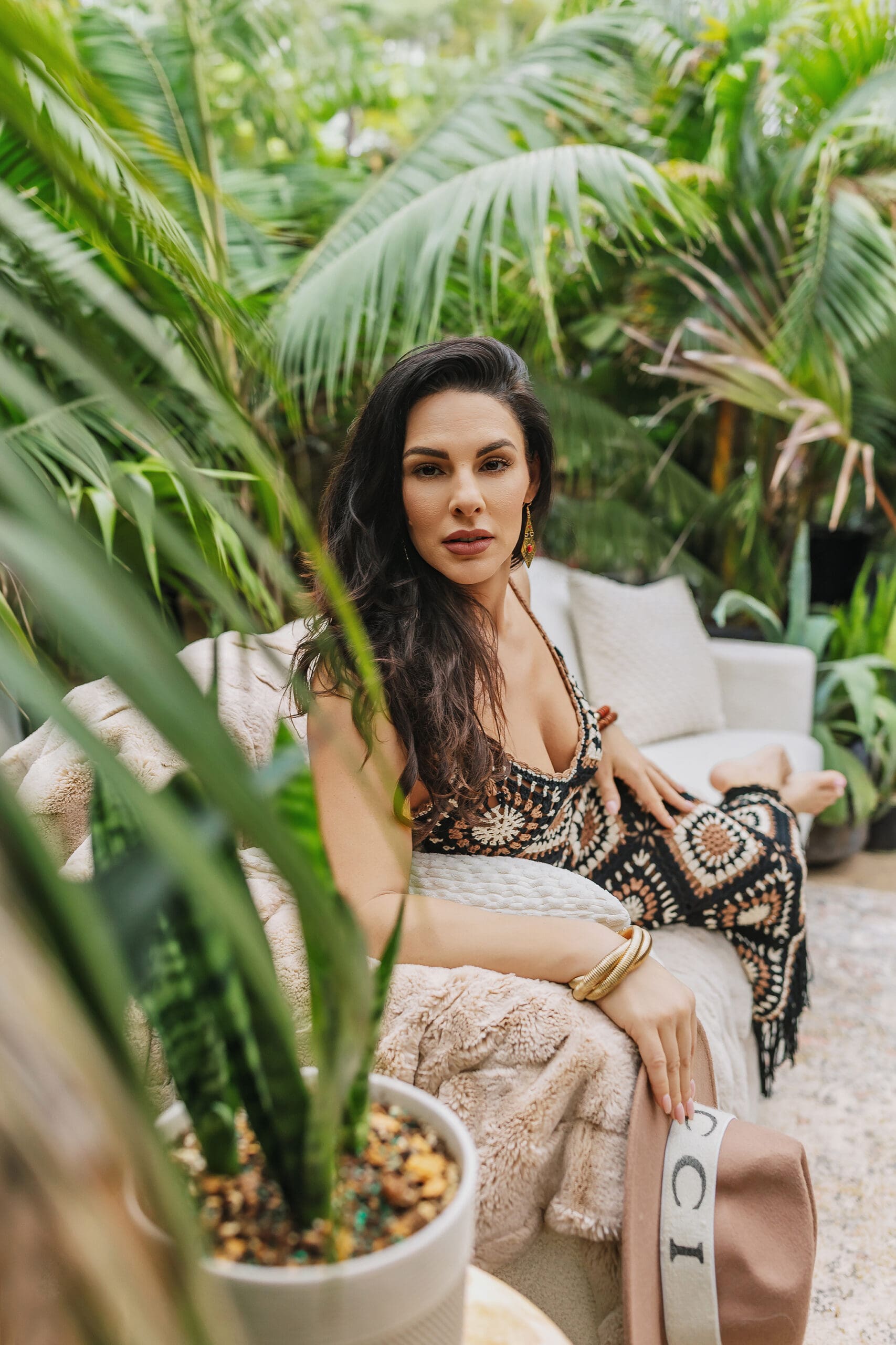 A woman with long dark hair in a patterned dress relaxes on an outdoor couch amidst lush green plants, exuding the tropical charm perfect for Connecticut branding photos. Holding a hat and adorned with gold earrings and bracelets, she epitomizes effortless elegance in an inviting setting.
