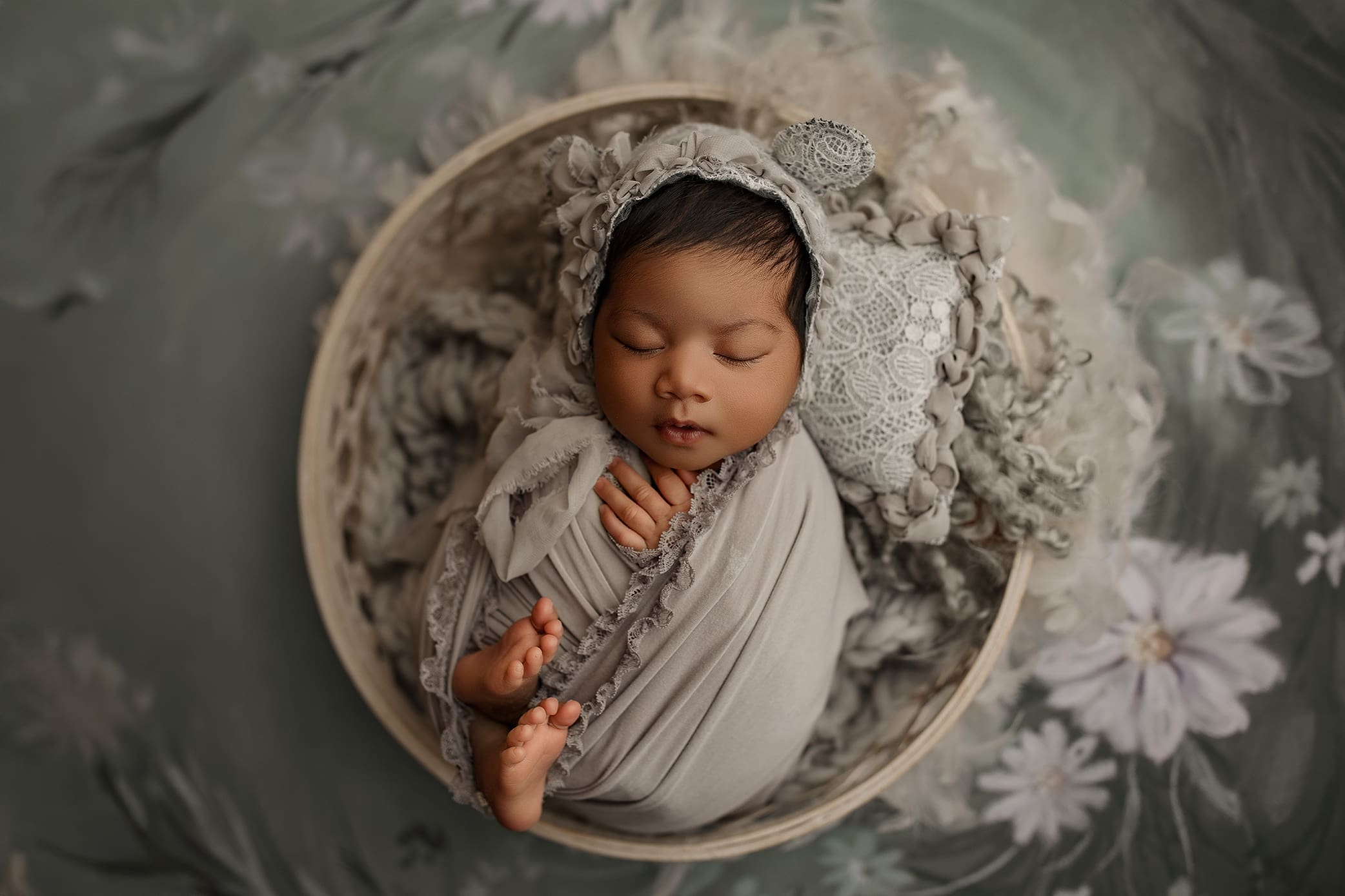 A sleeping baby, wrapped in a soft gray blanket with frilled edges, rests peacefully in a round basket, captured beautifully by a Connecticut newborn photographer. The baby wears a matching lace bonnet. The background features soft floral patterns in muted green and gray tones.