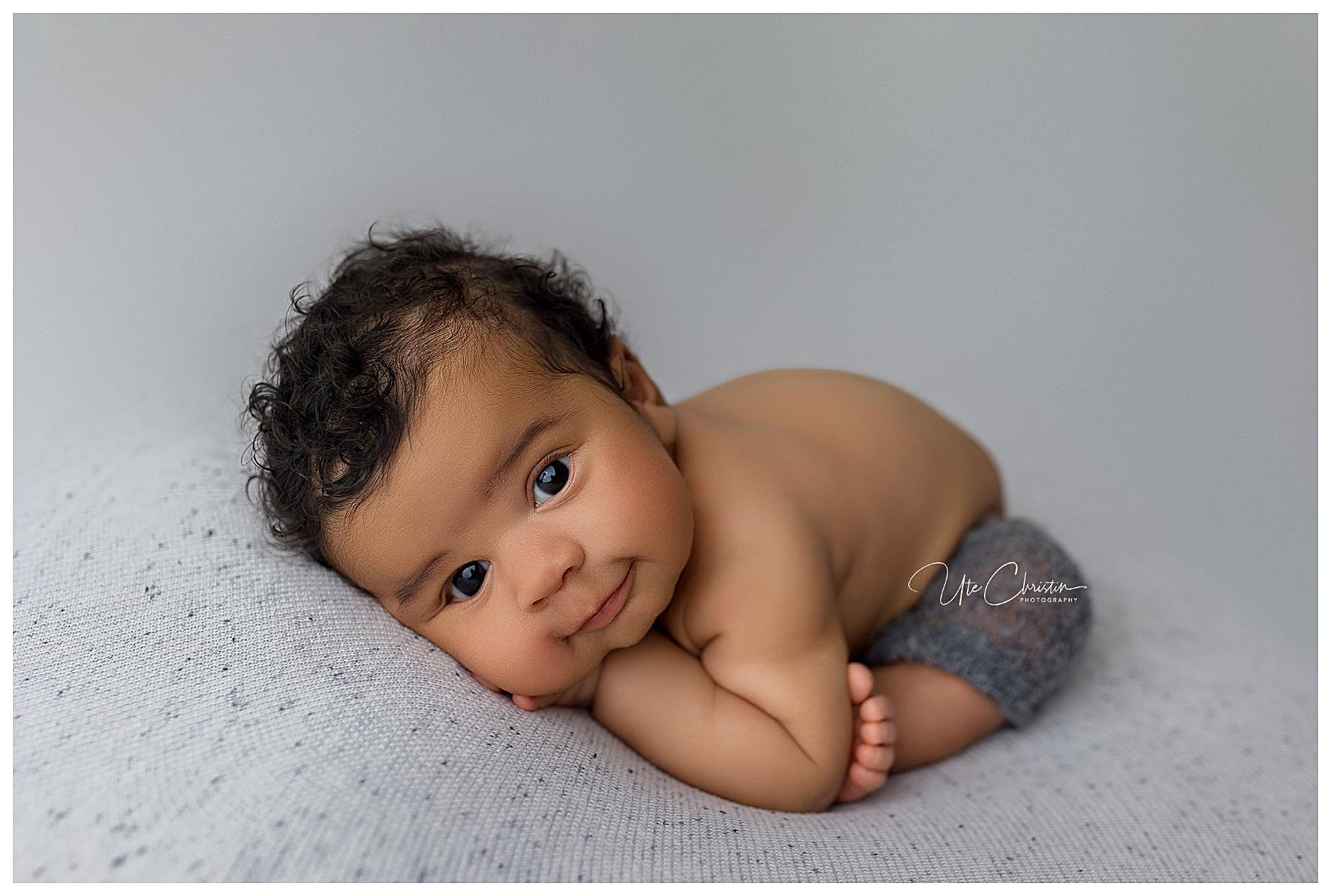 A baby with curly hair and brown eyes lies on their stomach, head resting on their arms. Dressed in grey shorts, they are on a textured light grey surface, looking directly at the camera with a gentle expression—just like those featured in baby stores in Connecticut.