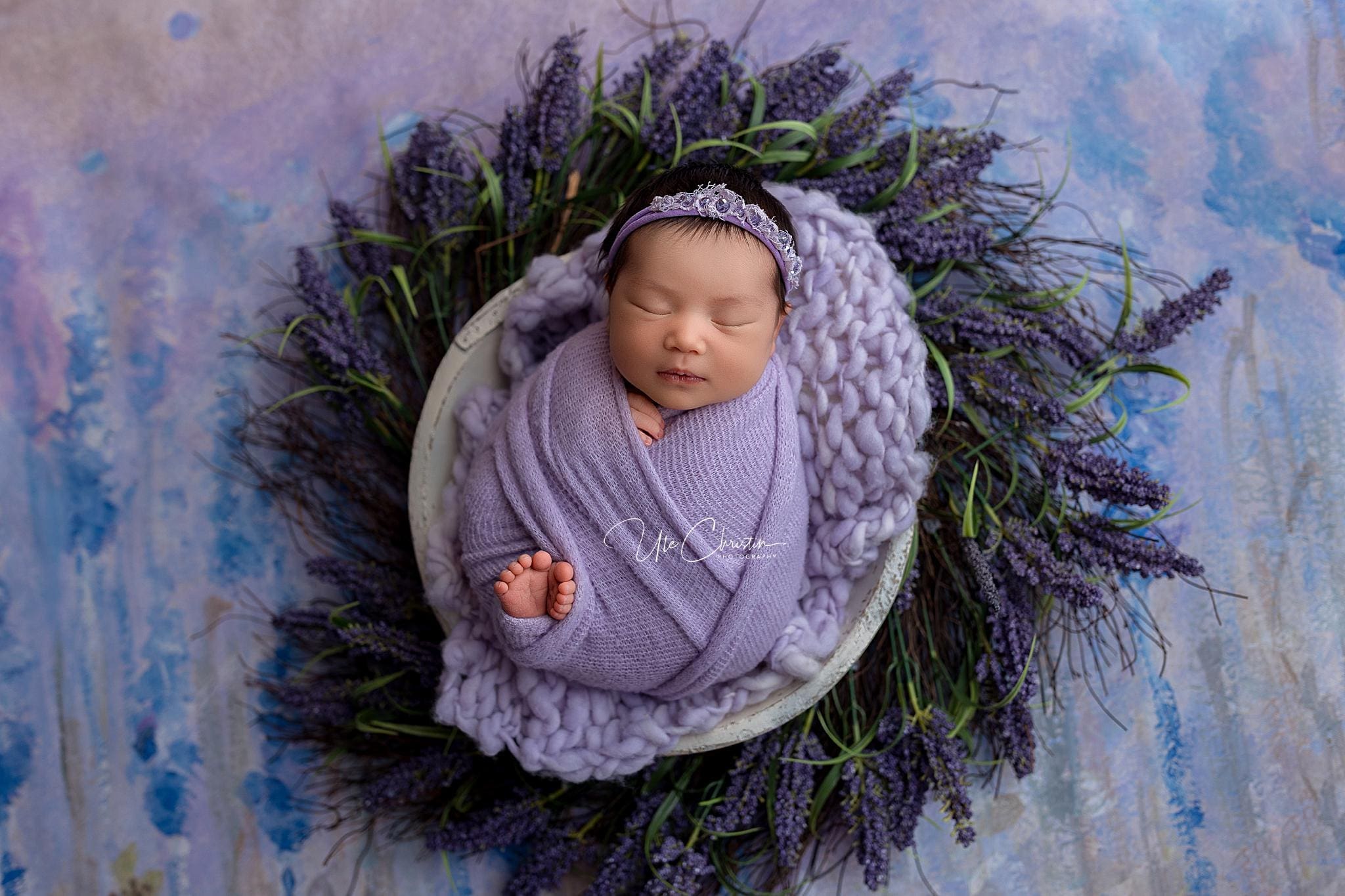 A newborn baby, peacefully sleeping and wrapped in a lavender blanket, wears a purple headband with a tiny tiara. The serene scene has been craftily set by one of the most caring doulas in CT, with the baby nestled among lavender flowers against a textured blue and purple backdrop.