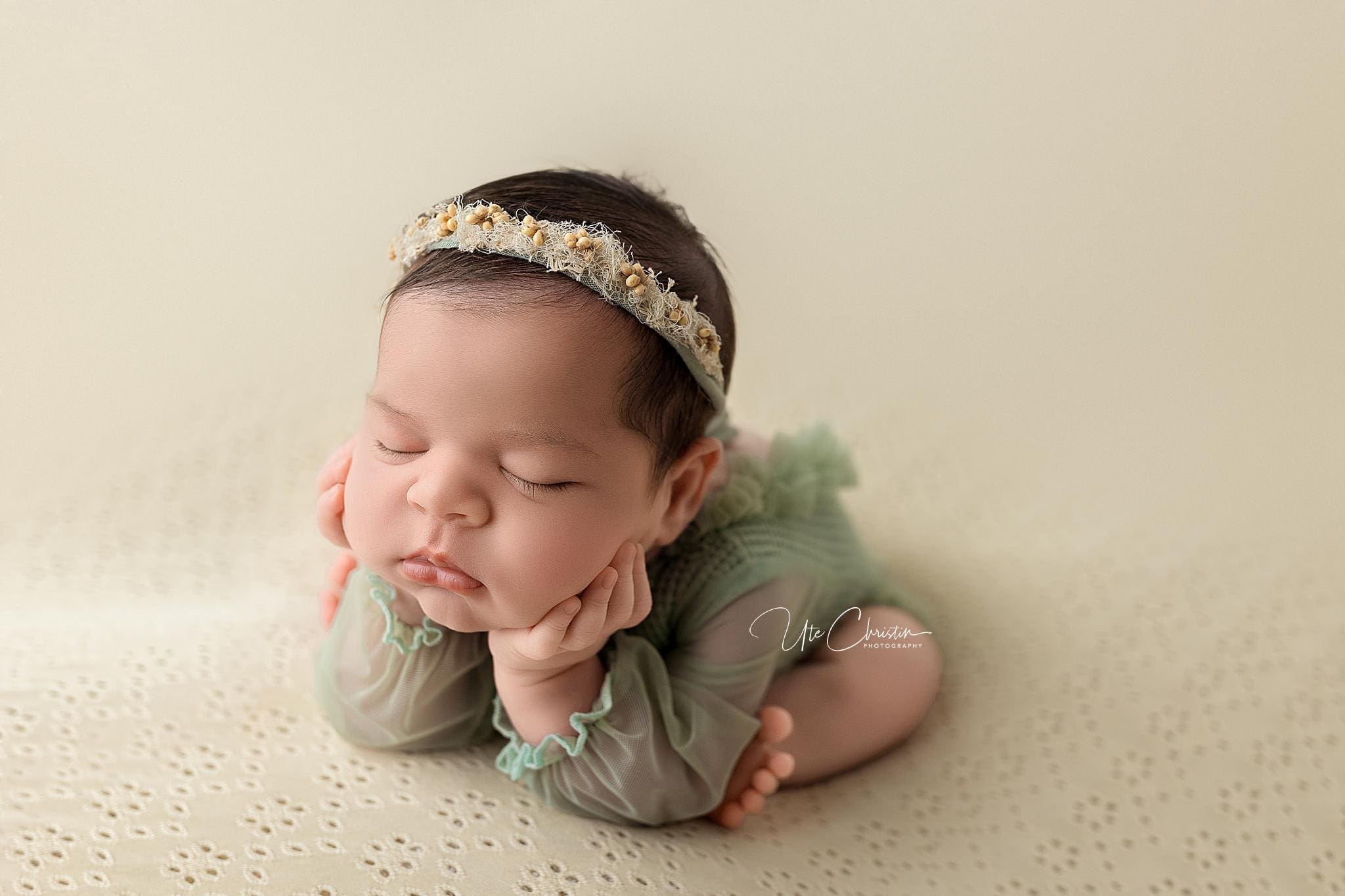 A sleeping baby wearing a floral headband and a green outfit, with their head resting on their hands. Midwives in Connecticut often capture these serene moments, showing the little one lying on a light-colored, textured surface.