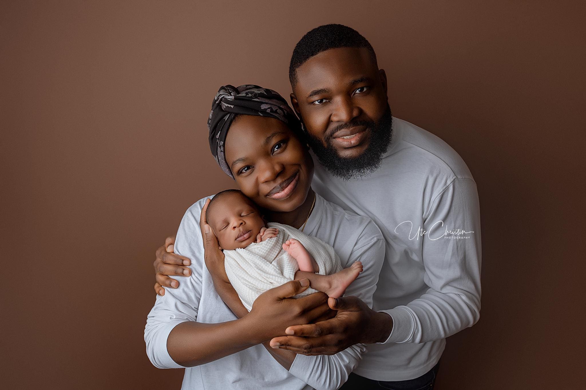 A joyful family portrait features a smiling couple holding a sleeping baby swaddled in white. They are all wearing white tops against a brown background, creating a cozy and harmonious atmosphere, reminiscent of the care and warmth provided by midwives in Connecticut.