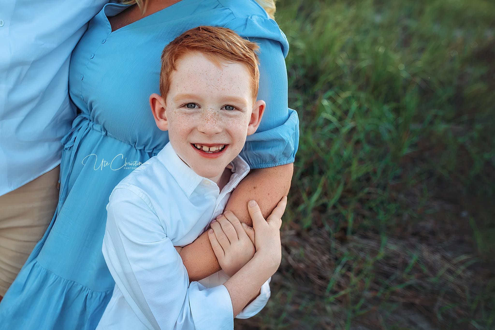 A smiling red-haired boy in a white shirt hugs the arm of a woman in a light blue dress, standing outside on green grass. The boy, looking directly at the camera, shows off his healthy smile—perfect for Pediatric Dentists In Milford CT.