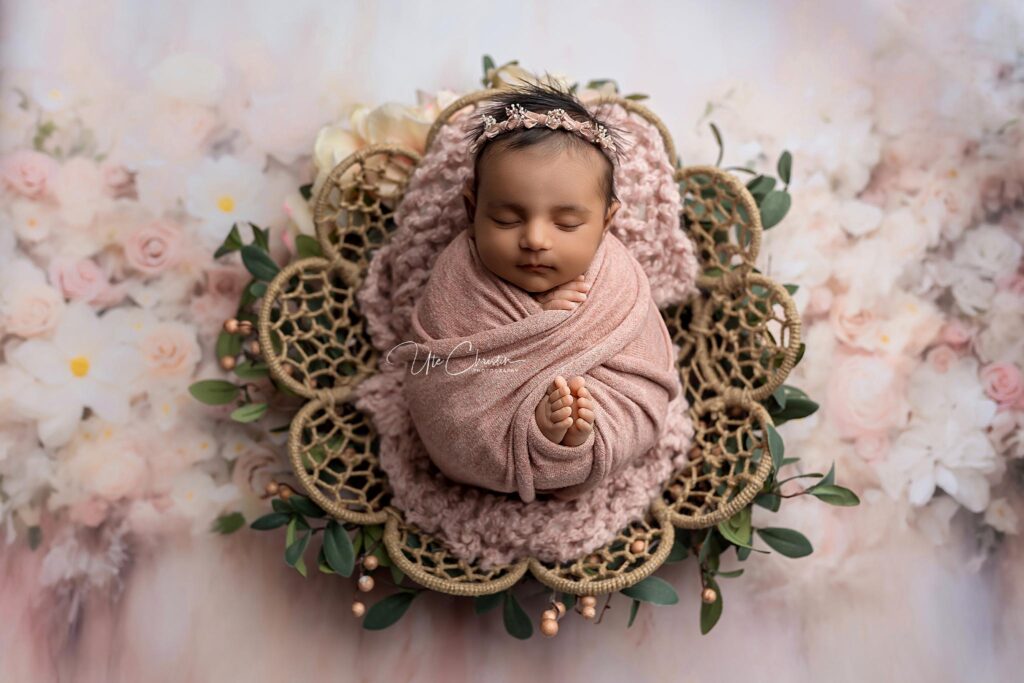 A newborn baby is peacefully sleeping, swaddled in a soft pink blanket with a floral headband, lying on a crocheted mat surrounded by greenery and flowers—perfect inspiration for Baby Shower Venues in Milford CT.