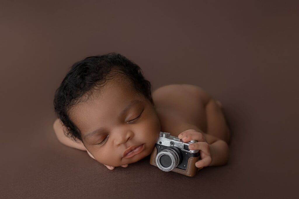 A sleeping newborn baby lies on a soft brown surface, holding a small vintage camera. With curly dark hair and a peaceful expression, the serene scene captures the essence of new beginnings—a moment cherished by parents and lactation consultants in CT alike.