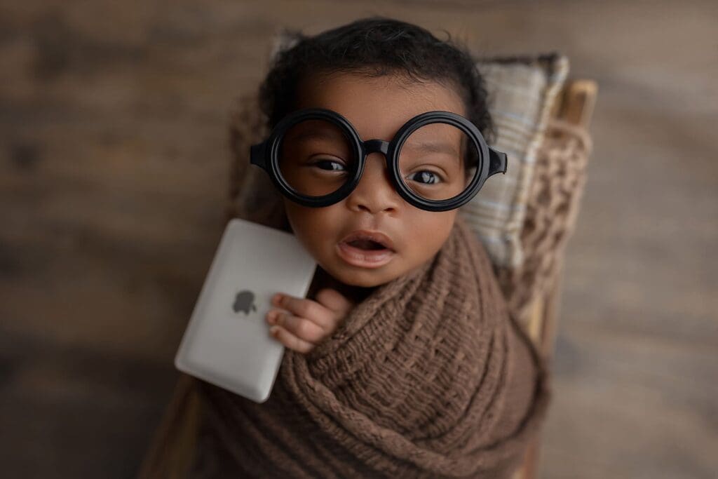 A baby wrapped in a brown blanket holds a smartphone and wears oversized round glasses, gazing up with a curious expression. Its as if the little one is already searching for lactation consultants in CT against the backdrop of a wooden surface.
