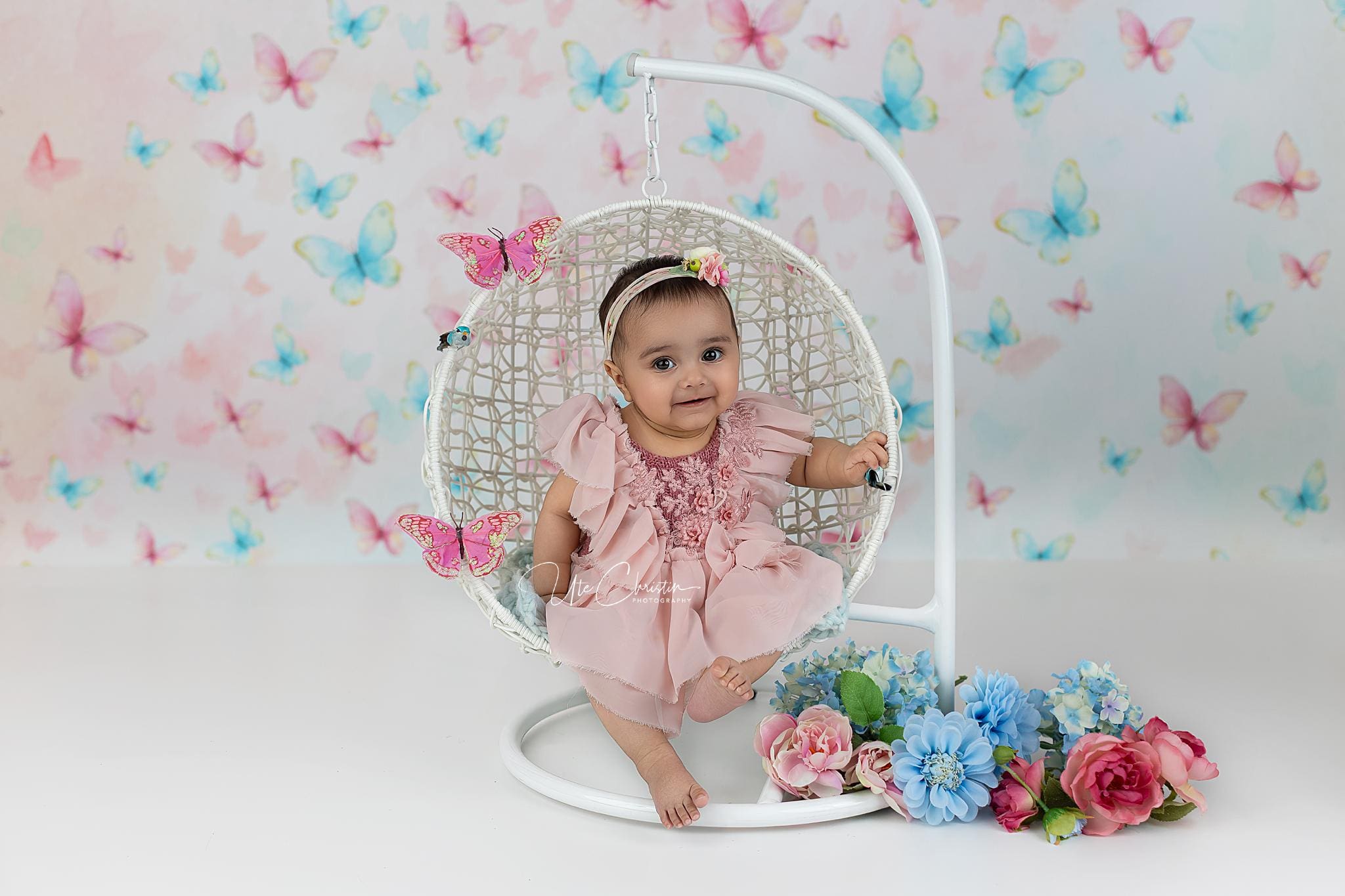 A smiling baby girl in a pink dress sits in a hanging white wicker chair decorated with pink butterflies. The scene, perfect for families and Nannies In Connecticut, features colorful butterflies and blue and pink flowers beside the chair.
