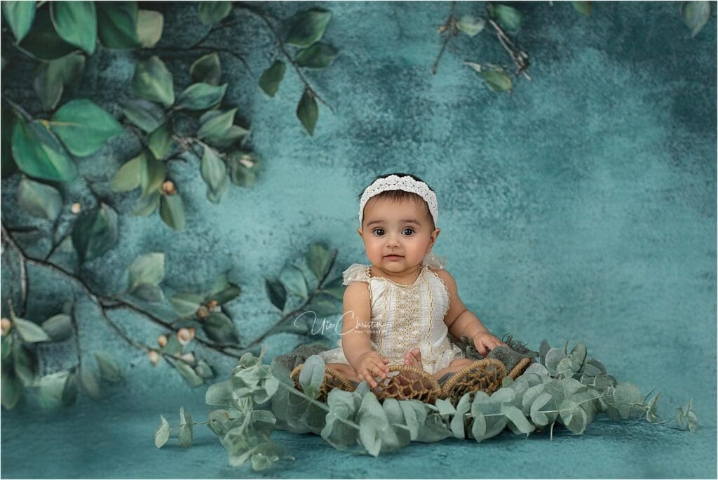 A baby wearing a white romper and headband sits on a woven mat surrounded by green leaves, with a teal background featuring leafy branches—a serene scene often cherished by nannies in Connecticut. The baby gazes calmly at the camera.