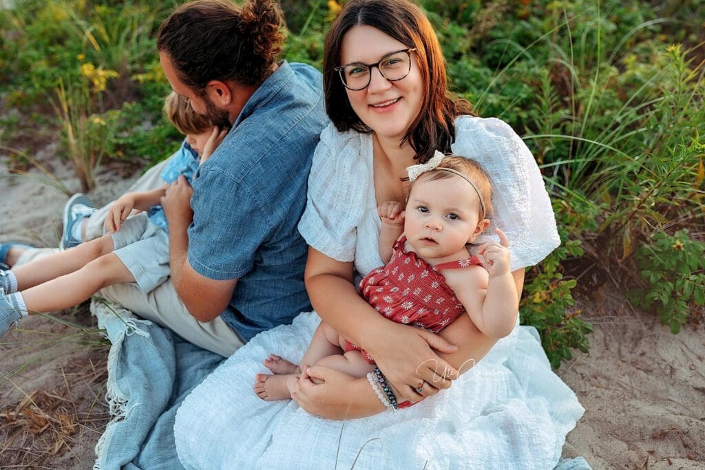 A woman in a white dress sits on a blanket outdoors, smiling at the camera while holding a baby girl in a red outfit. Nearby, a man and young boy sit with their backs turned—enjoying family time after visiting pediatricians in Milford CT.