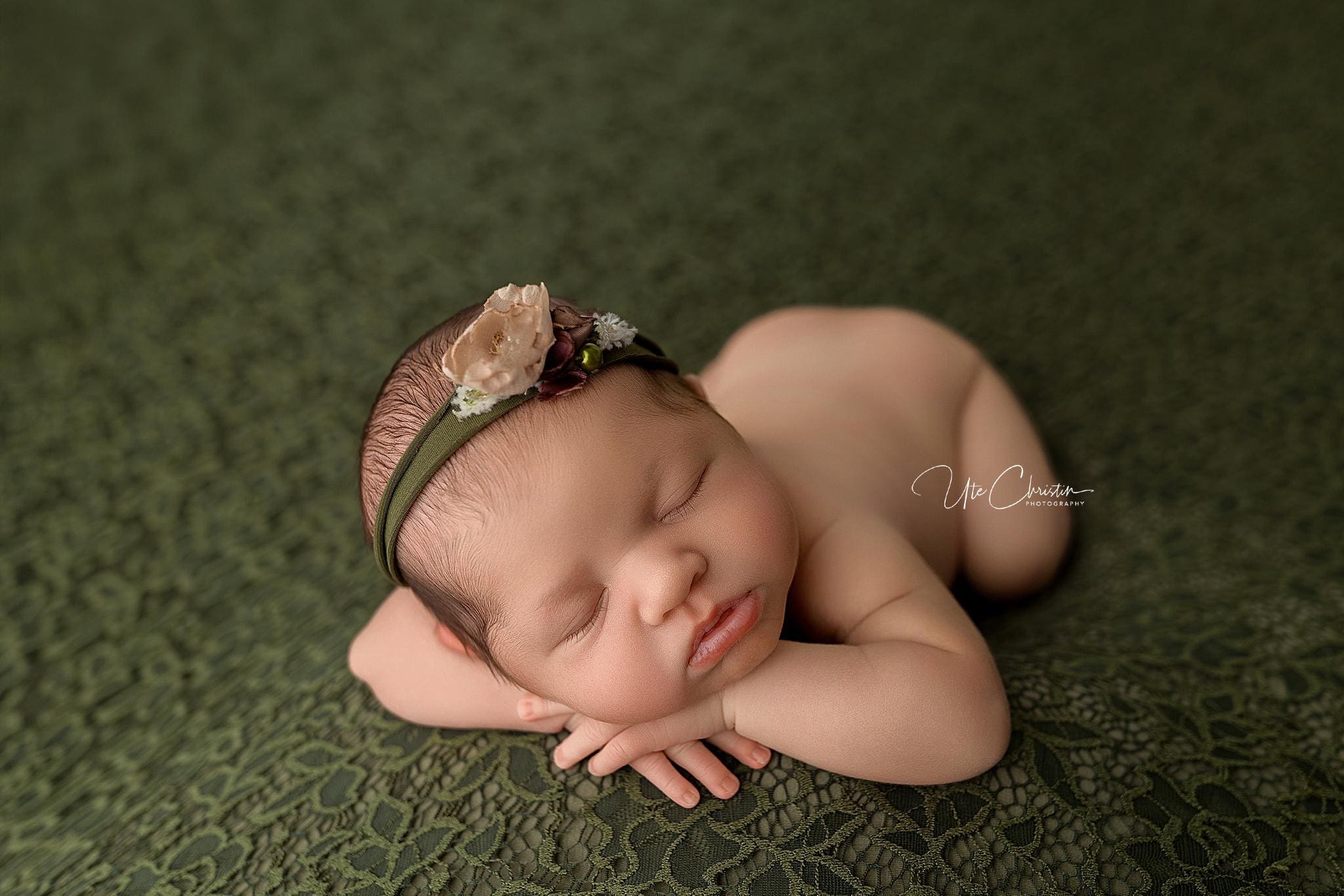 A newborn baby sleeps on a green lace blanket with arms folded under its head, wearing a floral headband with soft pink and green flowers—peaceful moments cherished by postpartum doulas in CT.