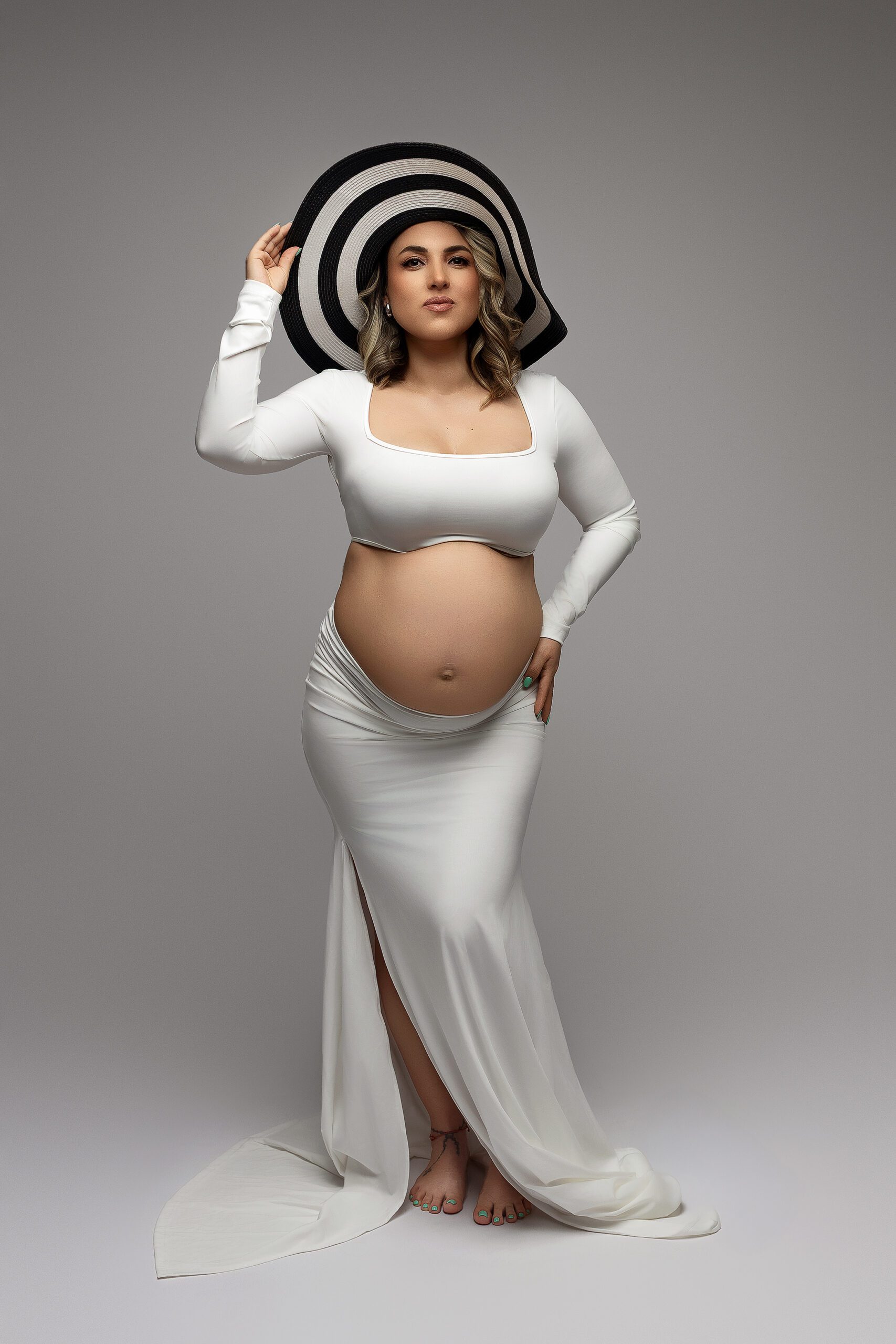 A pregnant woman in a white, long-sleeve crop top and skirt poses barefoot against a neutral background, holding a large black-and-white striped hat with one hand, revealing her baby bump during a maternity photoshoot in Connecticut.