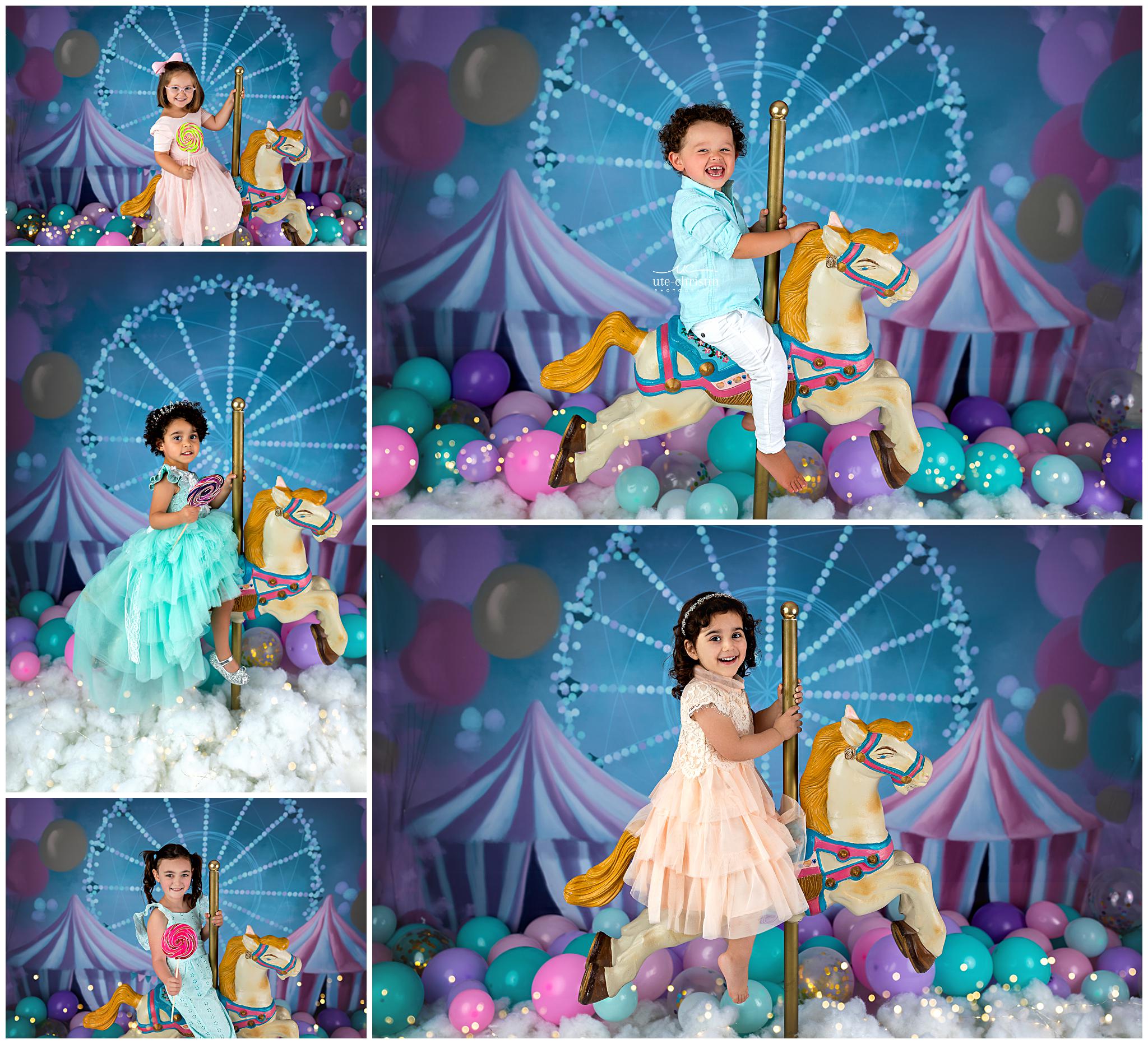 Four young children in colorful formal outfits pose on or beside a carousel horse, surrounded by balloons and pastel decorations with a Ferris wheel and circus tent backdrop, creating a festive, carnival atmosphere.