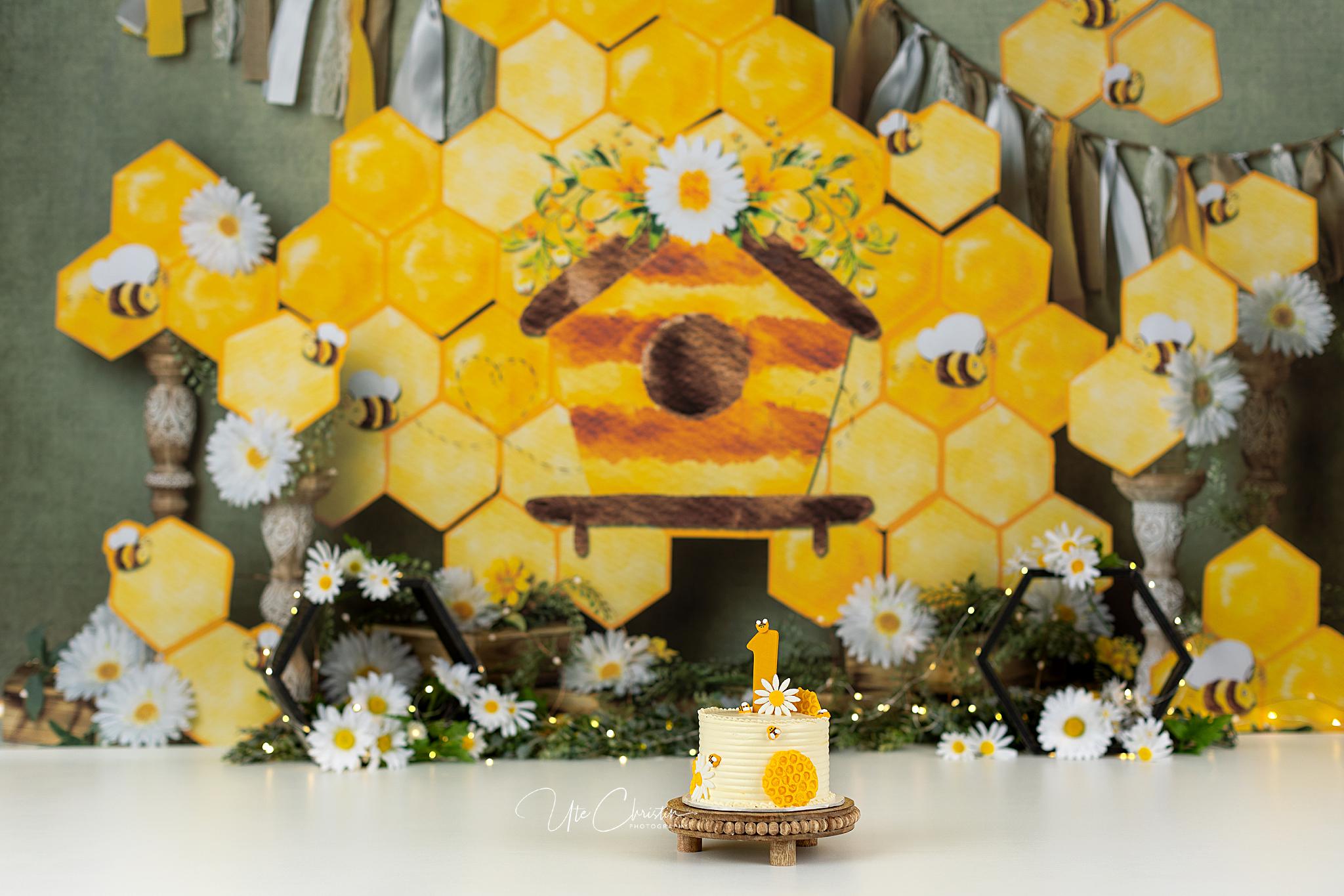 A small cake decorated with daisies and a honeycomb pattern sits on a stand. The background features a yellow beehive, honeycomb shapes, bees, and flowers, creating a bee and honey-themed setting.