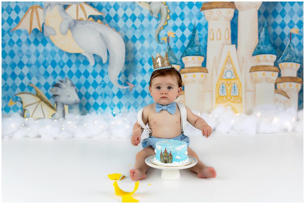 A baby wearing a crown, blue bowtie, and suspenders sits on the floor in front of a blue castle-themed cake. The background features drawings of a castle and dragons with a blue diamond pattern.