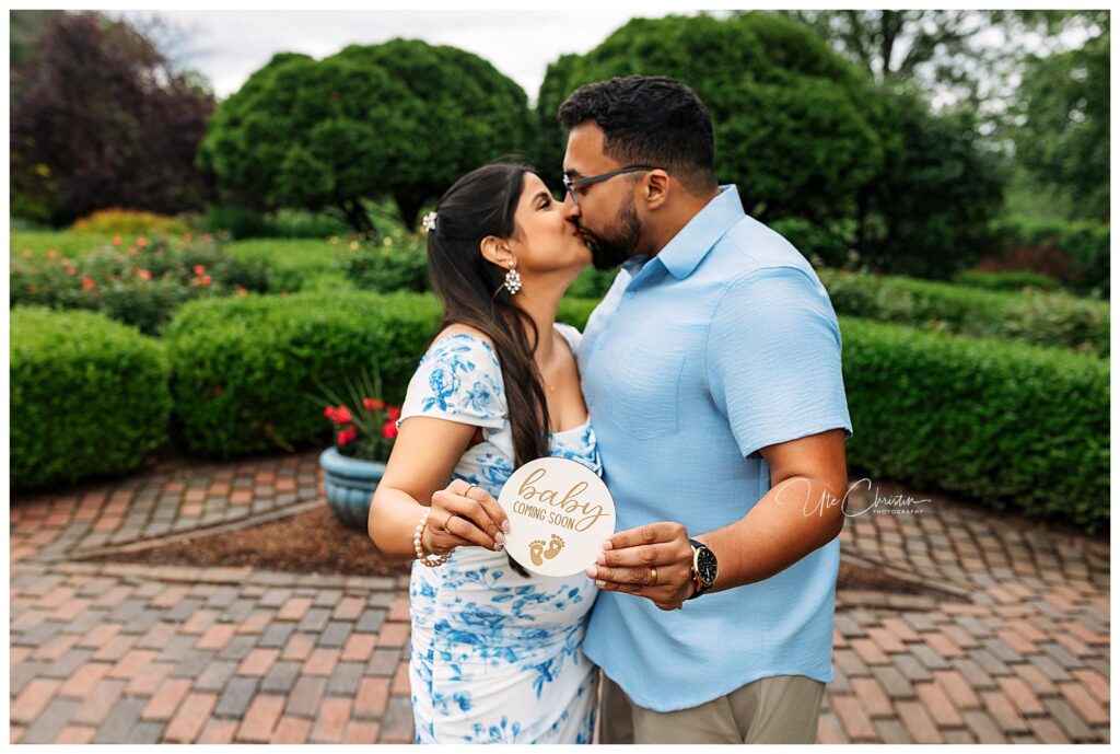 A couple stands in a garden, sharing a kiss. The woman, wearing a white and blue floral dress, holds a round sign that reads baby coming soon, indicating a pregnancy announcement. The man is in a light blue shirt.