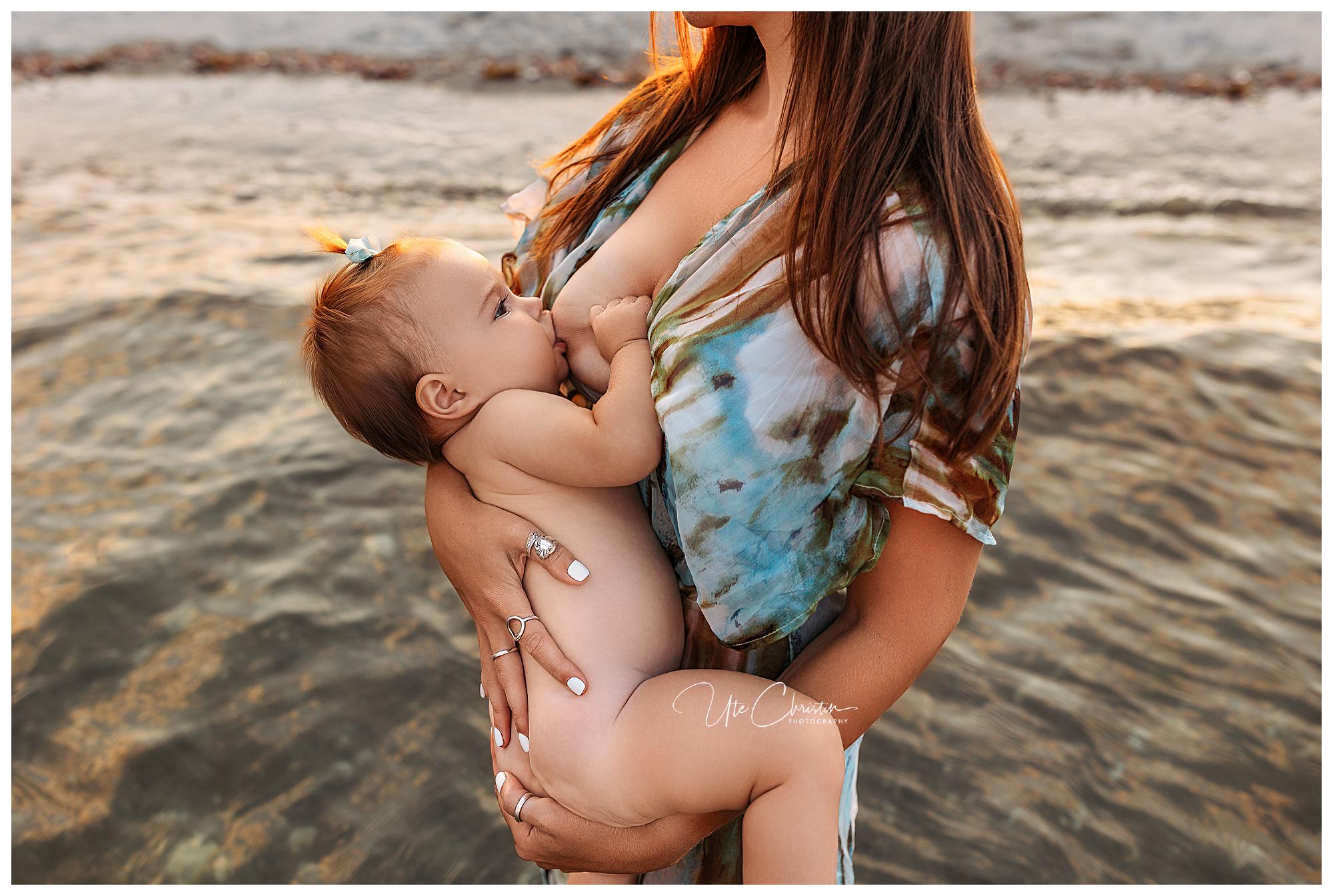 A woman in a patterned dress breastfeeds a baby while standing in shallow water at the beach during sunset. The babys head has a small bow and they are held securely in the woman's arms during their Connecticut breastfeeding session.
