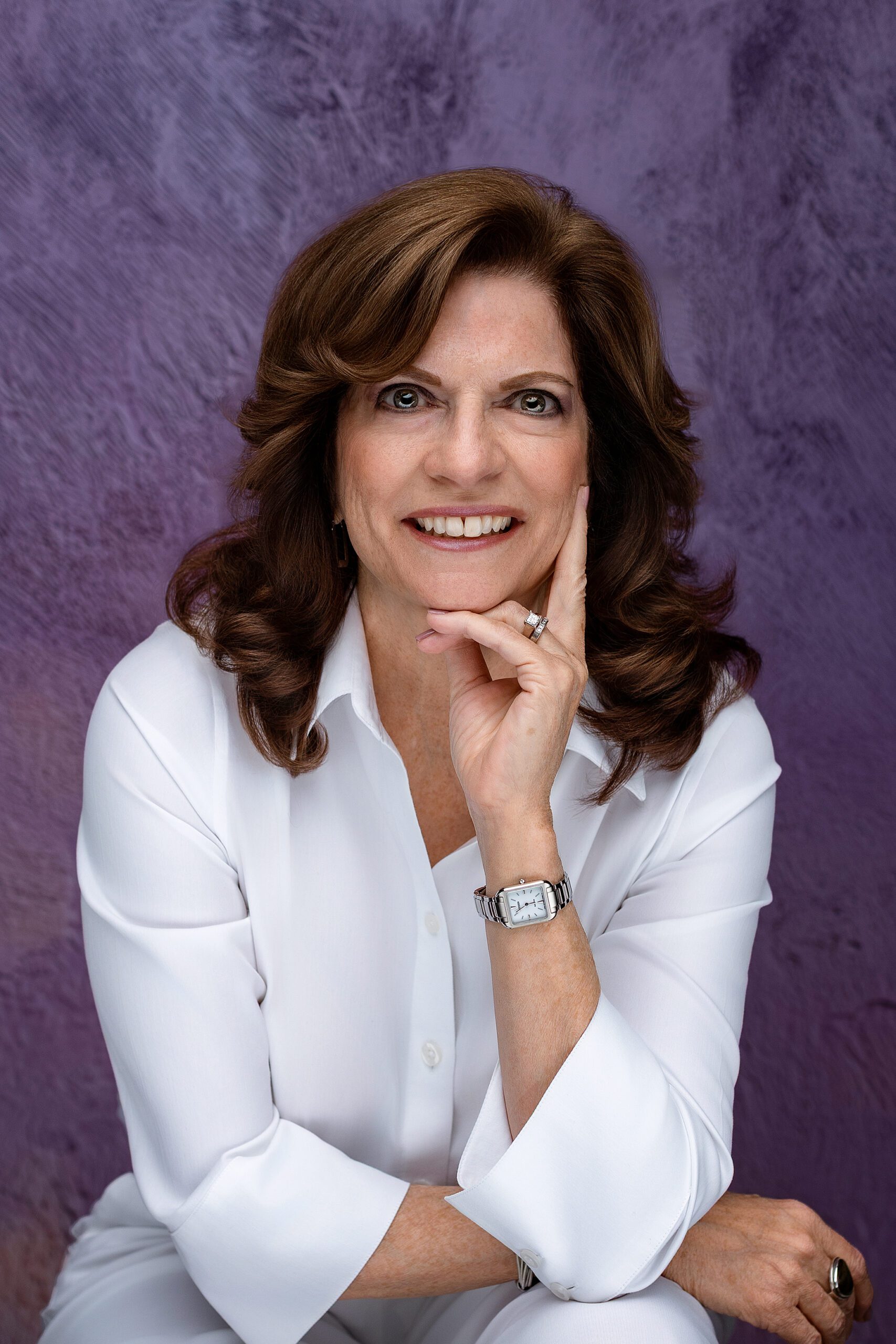 A woman with wavy brown hair, wearing a white shirt, silver watch, and ring, smiles softly while resting her chin on her hand, seated in front of a textured purple background for her CT headshot.